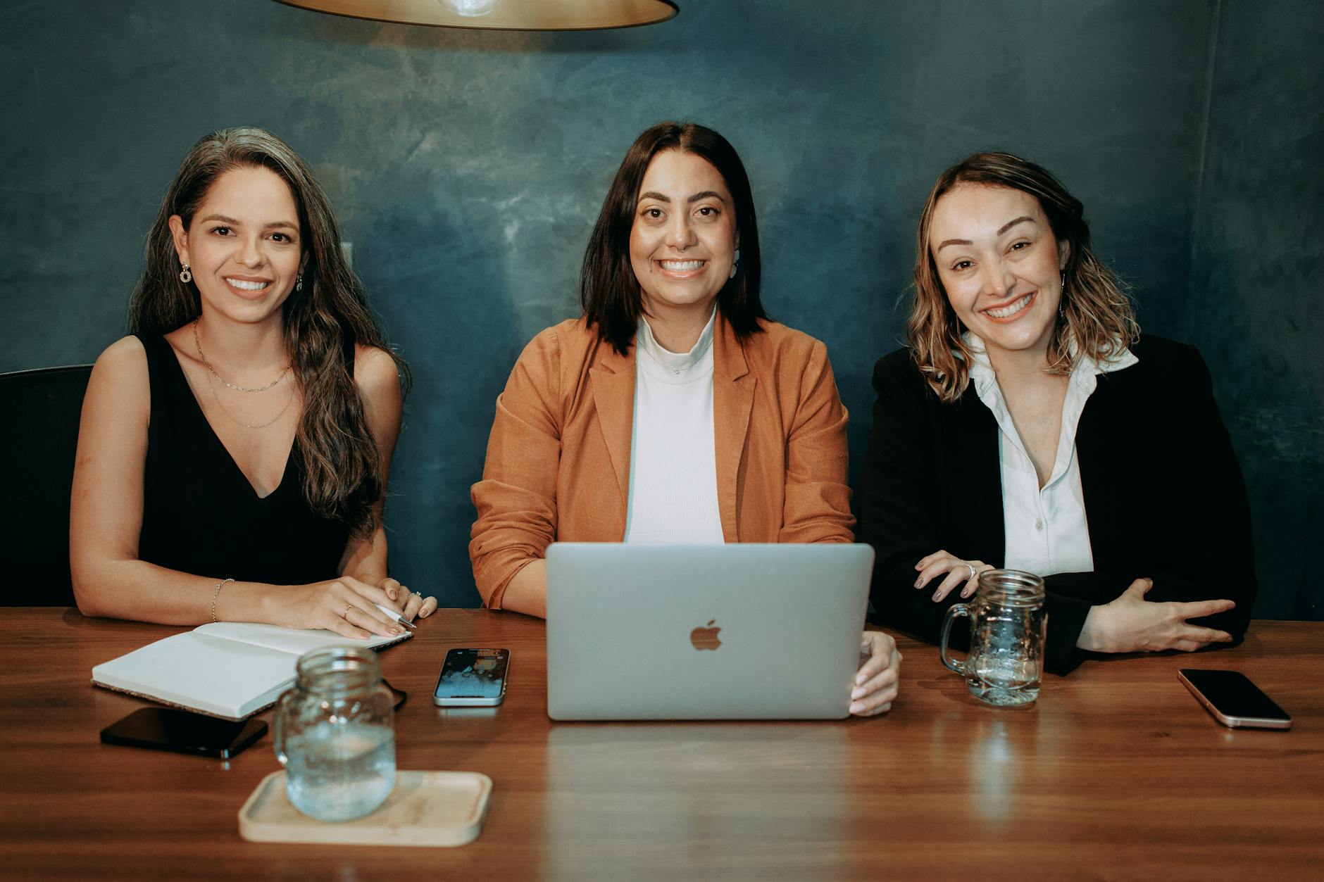Three professional women sitting at a wooden desk in an office with laptops and notebooks.